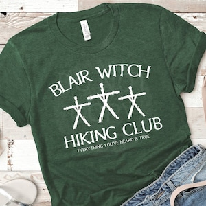 May include: A green t-shirt with white text that reads "Blair Witch Hiking Club" and "Everything you've heard is true". The shirt also features three stick figures with crosses.