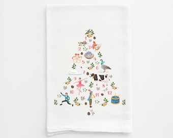 May include: White kitchen towel with a Christmas tree design featuring 12 illustrated images, including a bird, a nest, a goose, a cow, a ballerina, a drummer boy, and more. The numbers 1 through 12 are printed in red on the towel.