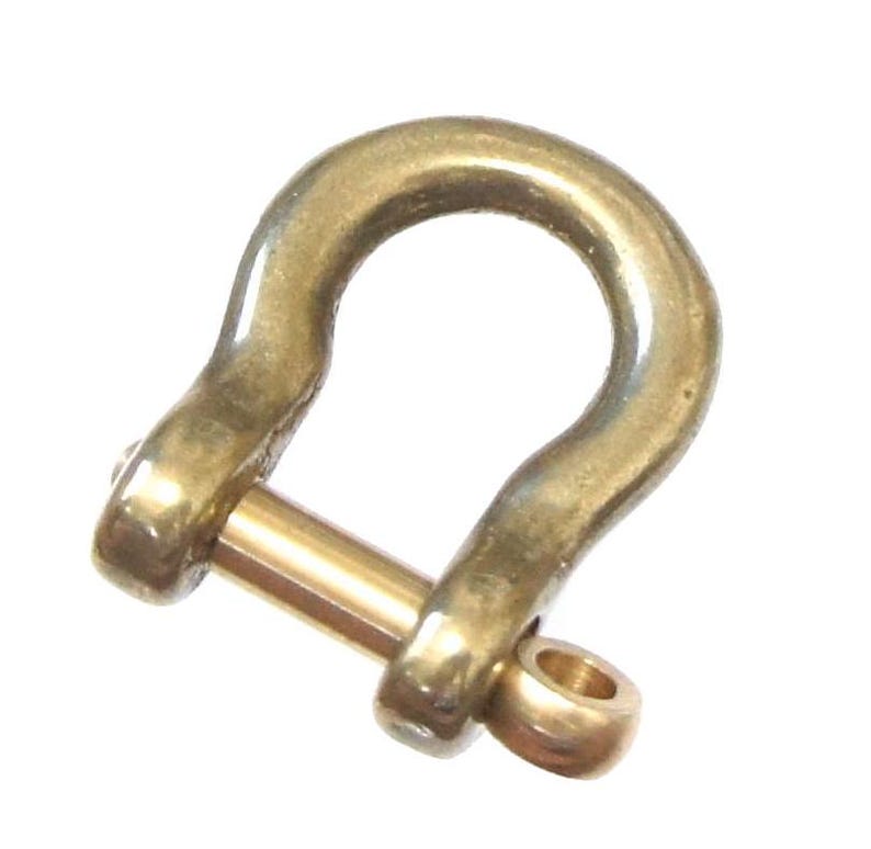 May include: A gold-colored metal shackle with a pin. The shackle is a U-shape with a rounded top and a pin that goes through the bottom of the U.