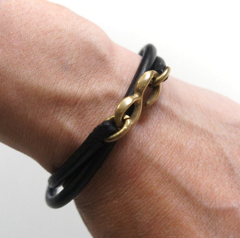 Puede incluir: Un brazalete de cuero negro con un cierre de s&iacute;mbolo de infinito dorado.