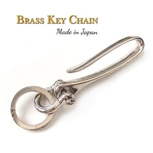 Könnte beinhalten: Ein silberfarbener Schlüsselanhänger aus Messing mit Haken und Ring. Der Text "Brass Key Chain" und "Made in Japan" sind in Gold bzw. Schwarz dargestellt.