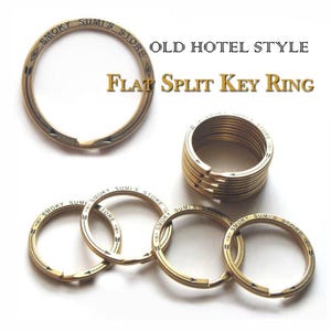 May include: A collection of brass-toned flat split key rings featuring the engraved text "SMOKY SUMI'S STORE" along the circumference. The rings are arranged individually and in a stacked formation, displaying a metallic finish and arrow motifs.