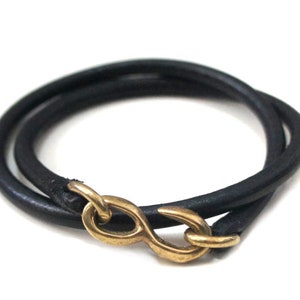 Puede incluir: Un brazalete de cuero negro con un cierre de oro en forma de infinito.