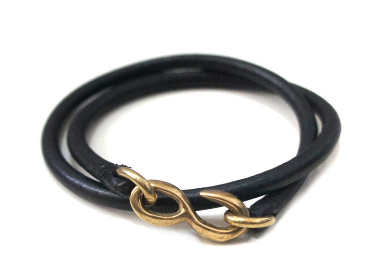 Puede incluir: Un brazalete de cuero negro con un cierre de oro en forma de infinito.