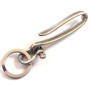 May include: A silver-colored metal keychain hook with a ring and a curved hook. The hook is designed to attach to a belt loop or bag, and the ring is for keys. The metal has a slightly aged appearance.
