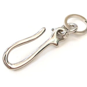 Op de afbeelding: Een zilveren sleutelhanger met een grote haak en een kleinere ring. De haak is gebogen en heeft een gladde afwerking.