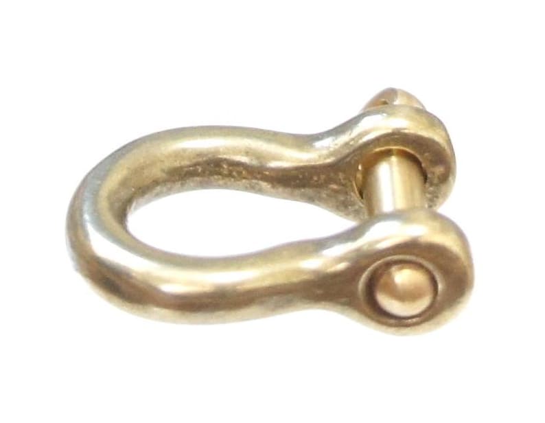 May include: A gold-colored metal shackle with a rounded, smooth surface and a pin through the center. The shackle is designed for attaching ropes or chains.