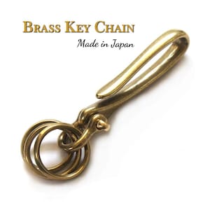 May include: A brass key chain with a hook and three rings. The text "Brass Key Chain Made in Japan" is in the upper portion of the image. The key chain is made of a shiny, gold-coloured metal.