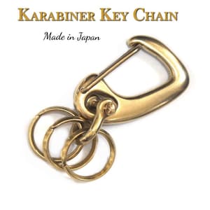 Könnte beinhalten: Ein goldfarbener Karabiner-Schlüsselanhänger aus Metall mit drei daran befestigten Schlüsselringen. Der Schlüsselanhänger trägt den Text "KARABINER KEY CHAIN" und "Made in Japan".
