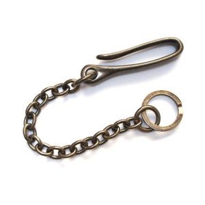 May include: A bronze-colored keychain with a hook, chain, and ring. The hook is curved and attached to a short chain of oval links. The chain connects to a small ring. The metal has a slightly aged appearance.