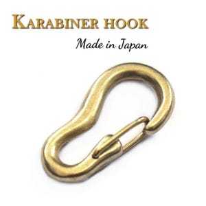 Op de afbeelding: Een gouden metalen karabijnhaak met een veerbelaste sluiting. De haak is gemarkeerd met "KARABINER HOOK Made in Japan".
