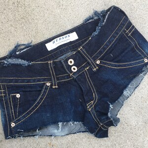 May include: Dark blue denim shorts with frayed edges and a small pocket on the front. The shorts have a label that reads "HUDSON -  jeans that fit you".