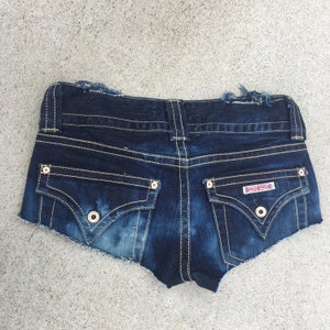May include: Dark blue denim shorts with frayed edges and a small red and white logo on the back pocket.