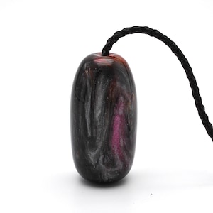 May include: A smooth, cylindrical pendant made of polished resin with a marbled pattern of black, silver, and deep magenta swirls. The pendant is suspended from a thin, black braided cord that enters through a hole at the top of the piece.