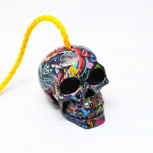 May include: A small, decorative skull with a vibrant graffiti-style design in various colors, including blue, red, yellow, and green. A yellow cord is attached to the top of the skull. The skull is set against a white background.