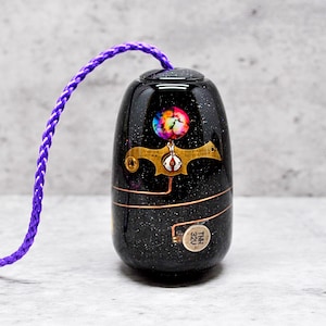 May include: A black and gold decorative bottle stopper with a galaxy design and a small clock face. The stopper has a purple cord attached to it.