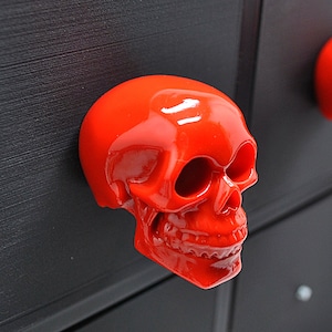 May include: A bright red skull-shaped drawer pull. The skull has a detailed face with a prominent nose and open mouth.