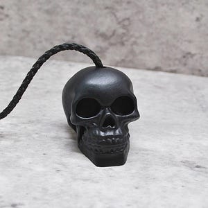 May include: A black skull-shaped pull for a light fixture. The skull is made of a smooth, matte black material and has a rope attached to the top.