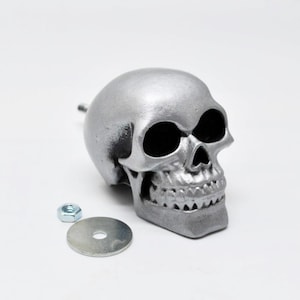 May include: A silver-colored skull-shaped decorative object with a detailed design, featuring a prominent jawline and eye sockets. Includes a washer and a nut. The skull is likely made of metal.
