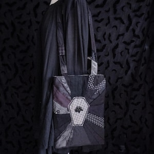 May include: A black tote bag featuring a central coffin-shaped patch with a small black floral emblem. The bag is constructed from various black fabric panels with different textures and patterns, including stripes and dots, and has two shoulder straps.
