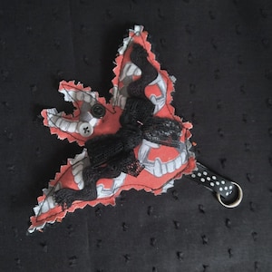 May include: A handmade, leaf-shaped fabric ornament with a red and gray pattern, black lace, and a small button. It has a black and white polka dot strap with a metal ring, set against a dark background.