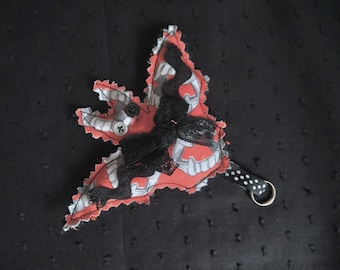 May include: A handmade, leaf-shaped fabric ornament with a red and gray pattern, black lace, and a small button. It has a black and white polka dot strap with a metal ring, set against a dark background.