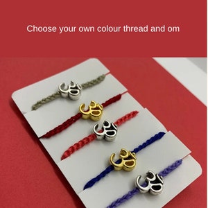 May include: Five Om bracelets with different coloured threads. The threads are grey, red, pink, blue, and purple. Each bracelet has a silver Om charm.