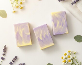 May include: Three rectangular bars of soap featuring a marbled pattern of soft lavender and pale yellow colors. The soap bars are arranged on a plain surface, surrounded by sprigs of purple lavender flowers, white chamomile flowers, and green leaves.