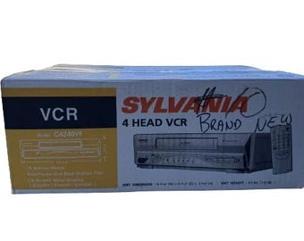 May include: A Sylvania 4 Head VCR in its original box. The box is light blue with black and yellow accents. The text "VCR" and "Brand New" are visible. The VCR has a remote control.