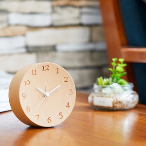 May include: A wooden clock with white hands and black numbers on a light wood surface. The clock is round and has a diameter of approximately 4 inches.