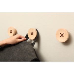 以下が含まれることがあります： 中央にXのデザインが施されたボタン型の木製壁掛けフックが3つ。黒いバッグがフックの1つにかけられています。