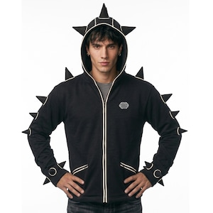 May include: Black zip-up hoodie with a distinctive design, featuring black spikes on the hood, shoulders, and sleeves. The hoodie has white trim and a small embroidered emblem on the chest. The model is wearing a grey shirt underneath.