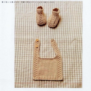 May include: A beige knitted baby bib and booties. The bib has a simple design with a button closure. The booties have a cable knit pattern. The items are displayed on a checkered fabric.