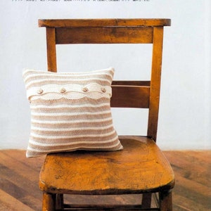 May include: A striped knitted cushion with a beige and white colour scheme. The cushion has a row of decorative buttons along the front. The cushion is sitting on a wooden chair.