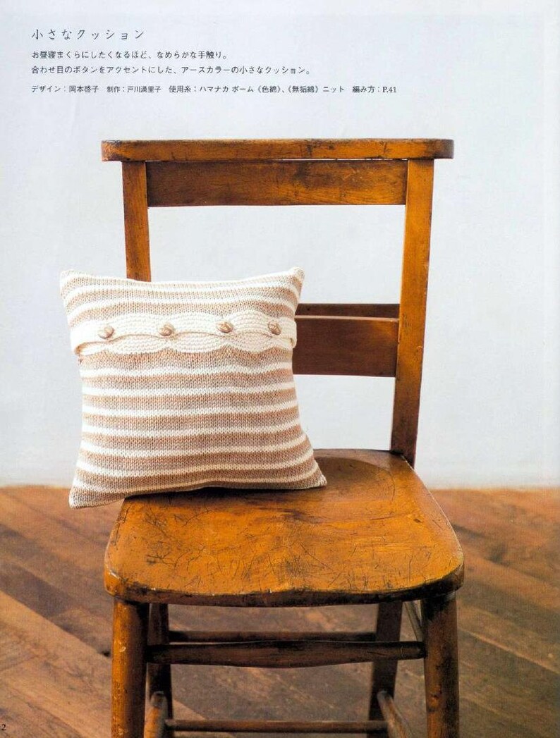 May include: A striped knitted cushion with a beige and white colour scheme. The cushion has a row of decorative buttons along the front. The cushion is sitting on a wooden chair.