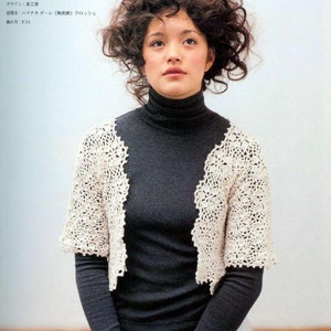 May include: A white crocheted bolero with a floral design. The bolero is worn over a black mock neck shirt and a white skirt.