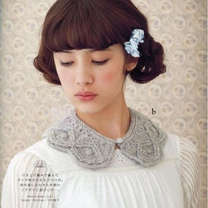 May include: A woman with brown hair and bangs is wearing a white blouse with a grey crocheted collar. The collar has a diamond pattern and a small button in the centre. The woman is wearing two blue flower hair clips.