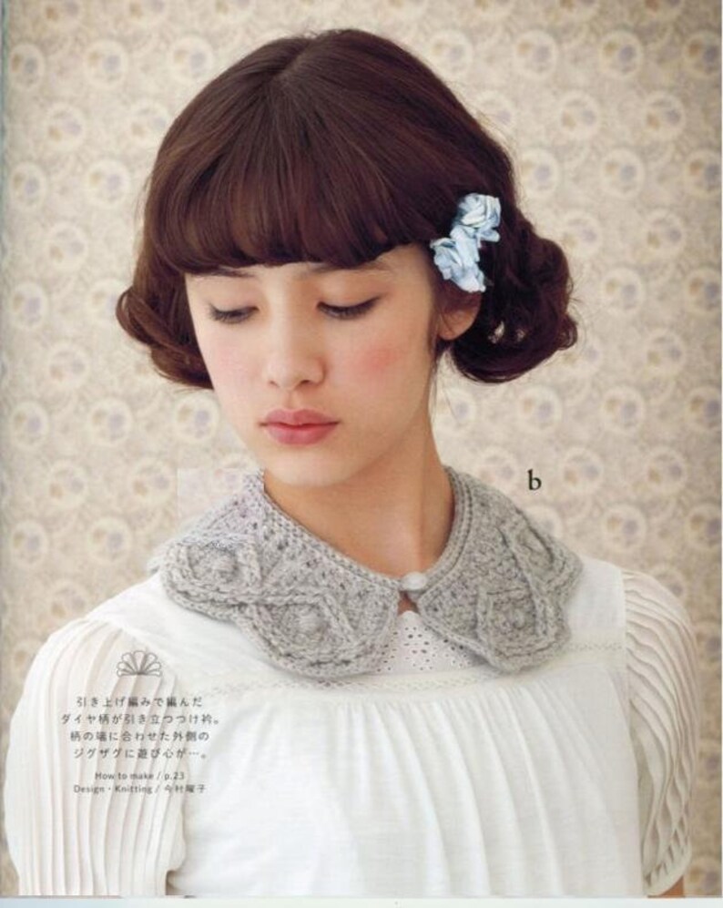 May include: A woman with brown hair and bangs is wearing a white blouse with a grey crocheted collar. The collar has a diamond pattern and a small button in the centre. The woman is wearing two blue flower hair clips.