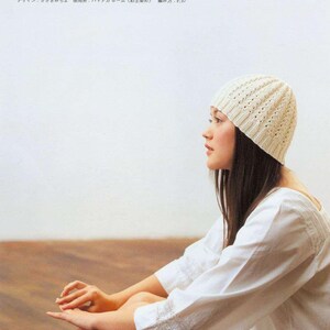 May include: A white crocheted beanie with a delicate openwork pattern. The beanie is worn by a person with long dark hair, sitting with their legs crossed.