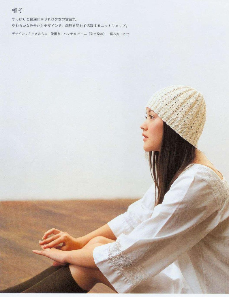 May include: A white crocheted beanie with a delicate openwork pattern. The beanie is worn by a person with long dark hair, sitting with their legs crossed.