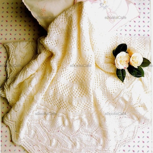 Puede incluir: Una manta de bebé de ganchillo blanco con un delicado patrón de encaje. La manta está doblada en una cama con una almohada blanca y una cinta rosa. Hay flores blancas sobre la manta.