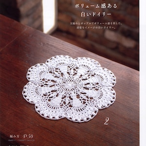 May include: A white crocheted doily with a delicate, lacy design. The doily is on a wooden surface. The text "ボリューム感ある 白いドイリー" is visible in the image.