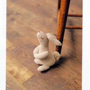 May include: A beige knitted rabbit amigurumi toy with a long, floppy ear, sitting on a wooden floor. The rabbit is looking up and has a sweet expression on its face.