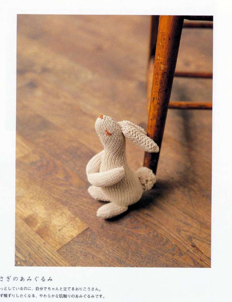 May include: A beige knitted rabbit amigurumi toy with a long, floppy ear, sitting on a wooden floor. The rabbit is looking up and has a sweet expression on its face.
