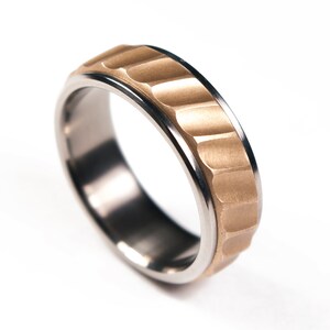 Può includere: Un anello in titanio con una fascia dorata che presenta un design testurizzato e rigato.