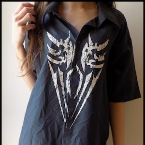 May include: Dark navy blue button-down shirt with a large white tribal design on the front. The short-sleeved shirt is worn over black fishnet stockings. The shirt has a classic collar and button closure.