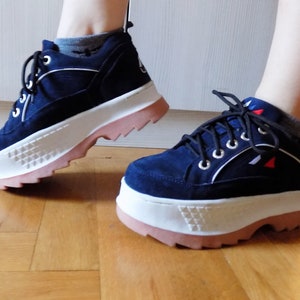 May include: Navy blue platform sneakers with white and pink accents. The shoes feature a suede-like upper, white soles with a geometric design, and pink rubber outsoles. The sneakers have black laces and silver eyelets.