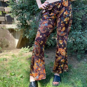 May include: Brown velvet bell-bottom pants with a colorful paisley pattern. The pants feature a high waist and a flared leg. The paisley design includes shades of blue, yellow, and orange. The pants are worn with black sandals.