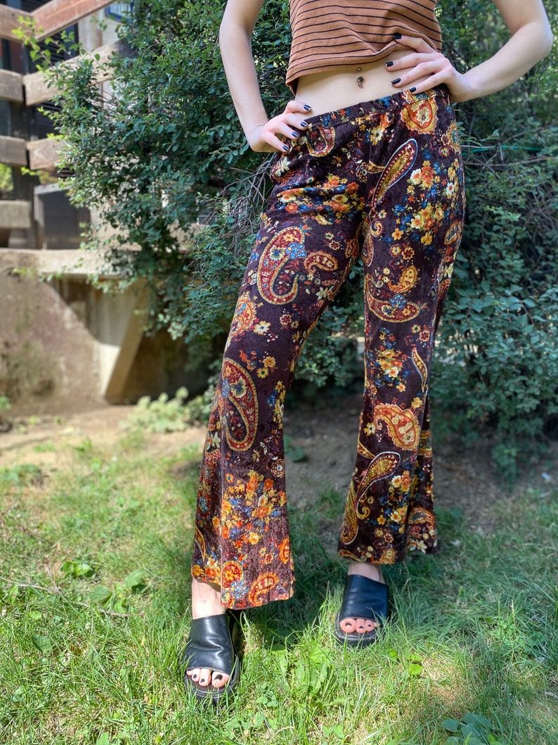 May include: Brown velvet bell-bottom pants with a colorful paisley pattern. The pants feature a high waist and a flared leg. The paisley design includes shades of blue, yellow, and orange. The pants are worn with black sandals.