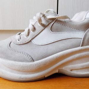 May include: A light gray platform sneaker with white accents. The shoe features a suede upper, a thick, wavy white sole, and white laces. The design includes a white side panel and a padded collar.